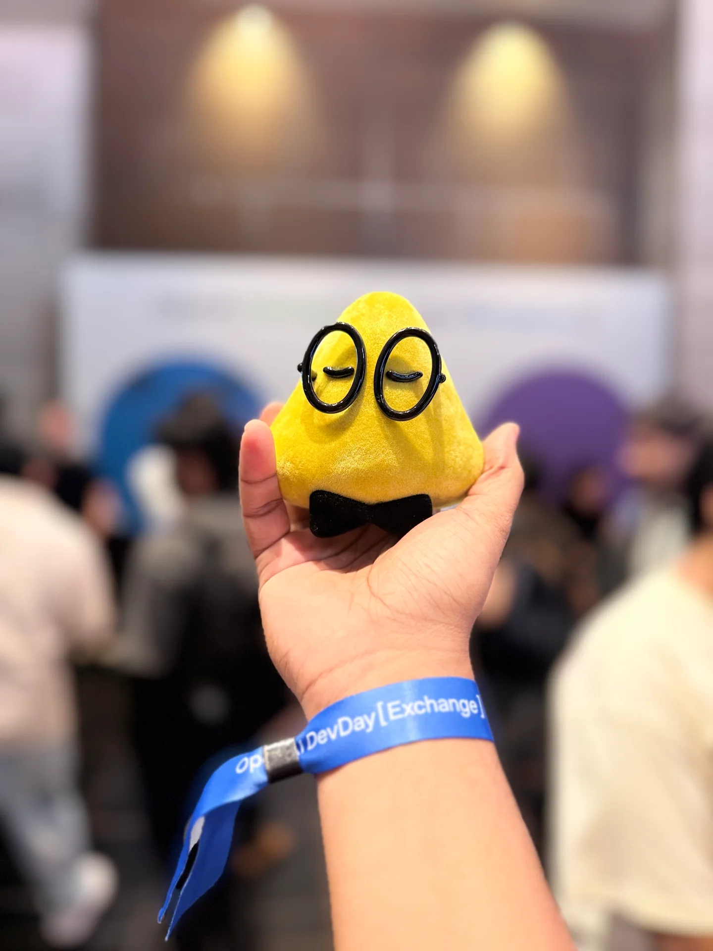 A yellow OpenAI builder mascot held above Robin Francis's DevDay Exchange wristband
